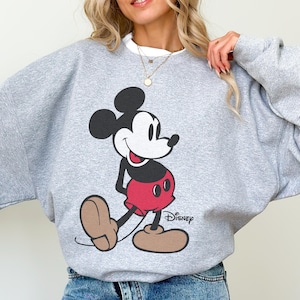 May include: A gray sweatshirt featuring a large graphic of Mickey Mouse in his classic pose. Mickey is depicted in black, red, and tan colors. The Disney logo is printed below the character.