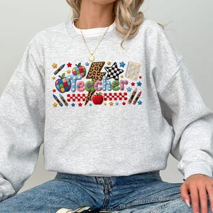 Puede incluir: Una sudadera gris claro de cuello redondo con un gr&aacute;fico colorido en el pecho que muestra la palabra Teacher en letras multicolores. El dise&ntilde;o incluye un rayo con estampado de leopardo, manzanas rojas, l&aacute;pices de colores, estrellas y un patr&oacute;n de estrella a cuadros.
