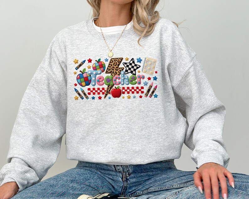 Puede incluir: Una sudadera gris claro de cuello redondo con un gr&aacute;fico colorido en el pecho que muestra la palabra Teacher en letras multicolores. El dise&ntilde;o incluye un rayo con estampado de leopardo, manzanas rojas, l&aacute;pices de colores, estrellas y un patr&oacute;n de estrella a cuadros.