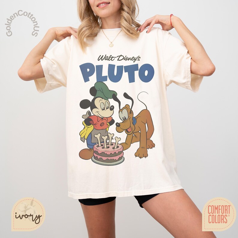 Puede incluir: Camiseta de color marfil con un dise&ntilde;o de Mickey Mouse y Pluto con una tarta de cumplea&ntilde;os. El texto "Walt Disney's PLUTO" est&aacute; impreso en azul. La camiseta es de la marca Comfort Colors.