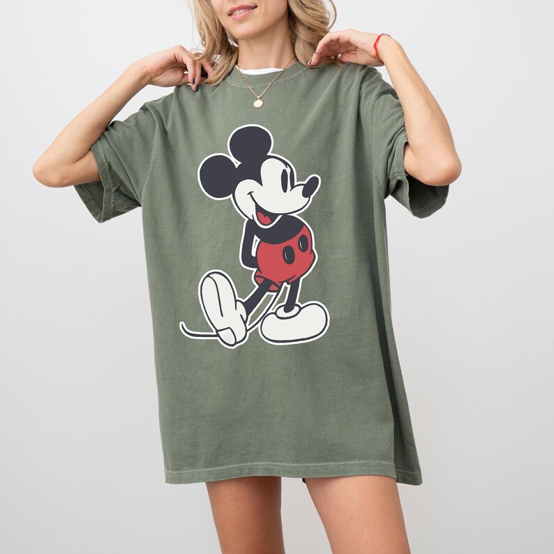 May include: Olive green t-shirt featuring a classic Mickey Mouse graphic. The cartoon character is depicted in black, red, and white. The shirt has short sleeves and a relaxed fit, suitable for casual wear.