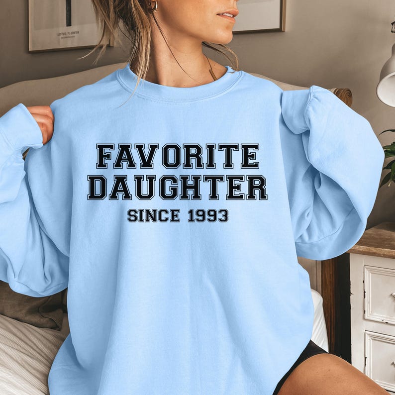 Peut inclure: Un sweat-shirt bleu clair avec l'inscription "FAVORITE DAUGHTER SINCE 1993" en lettres noires. Le sweat-shirt est de style col rond et semble &ecirc;tre fait d'une mati&egrave;re douce.