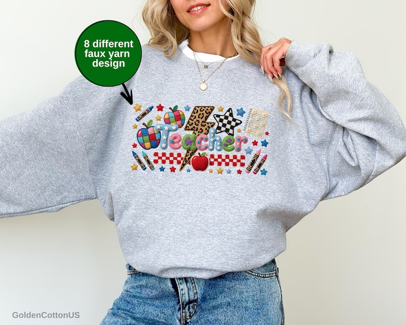 Puede incluir: Una sudadera gris claro de cuello redondo con un gr&aacute;fico colorido que muestra la palabra Teacher en un estilo de hilo falso texturizado. El dise&ntilde;o incluye un rayo con estampado de leopardo, manzanas, l&aacute;pices de colores, estrellas y un patr&oacute;n de cuadros. Un c&iacute;rculo verde con el texto 8 different faux yarn design se&ntilde;ala el gr&aacute;fico.