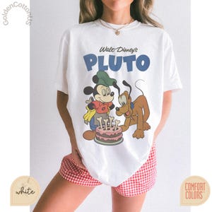 Puede incluir: Camiseta blanca con un gr&aacute;fico de Mickey Mouse y Pluto con una tarta de cumplea&ntilde;os. El texto "Walt Disney's PLUTO" es azul. La camiseta es de la marca Comfort Colors.