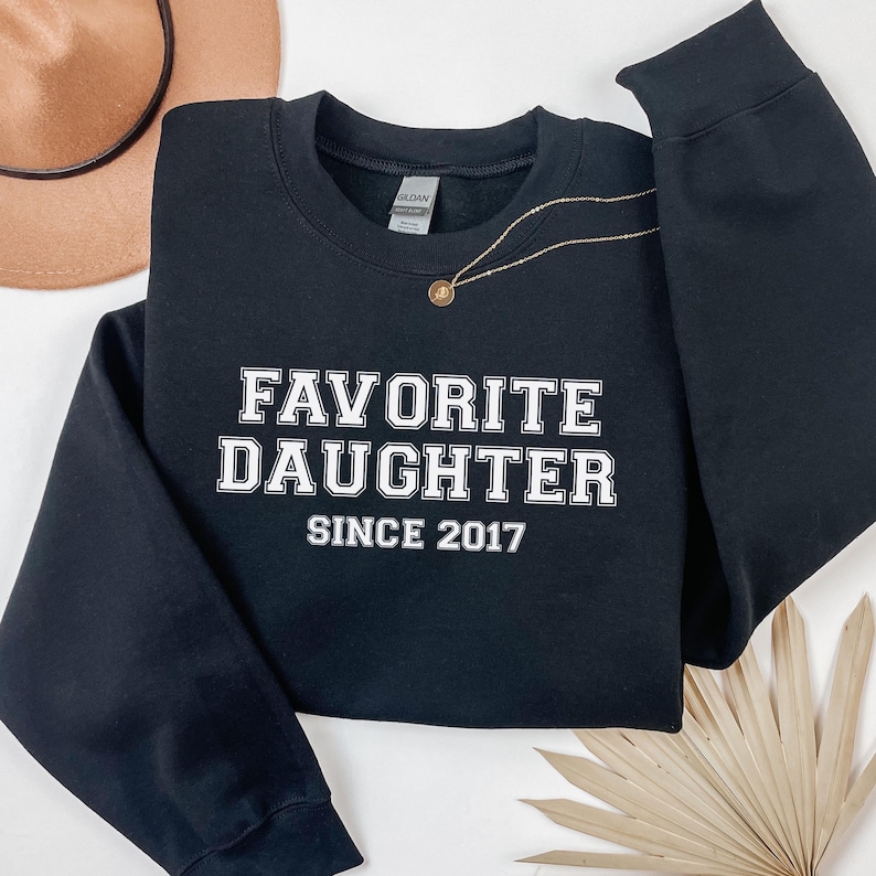 Peut inclure: Sweat-shirt noir avec l'inscription blanche "FAVORITE DAUGHTER SINCE 2017". Un collier dor&eacute; avec un pendentif est pos&eacute; sur le sweat-shirt. Un chapeau beige est visible en arri&egrave;re-plan.