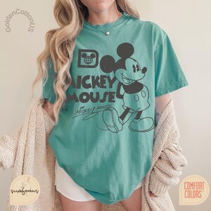 Puede incluir: Camiseta de color turquesa con un gr&aacute;fico negro de Mickey Mouse y el texto "Mickey Mouse". La camiseta tiene un aspecto vintage y cuello redondo. La camiseta est&aacute; hecha de un material suave y tiene mangas cortas. La camiseta es de Comfort Colors.