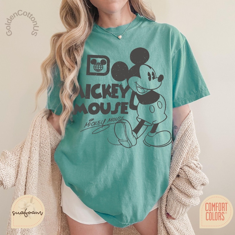 Puede incluir: Camiseta de color turquesa con un gr&aacute;fico negro de Mickey Mouse y el texto "Mickey Mouse". La camiseta tiene un aspecto vintage y cuello redondo. La camiseta est&aacute; hecha de un material suave y tiene mangas cortas. La camiseta es de Comfort Colors.