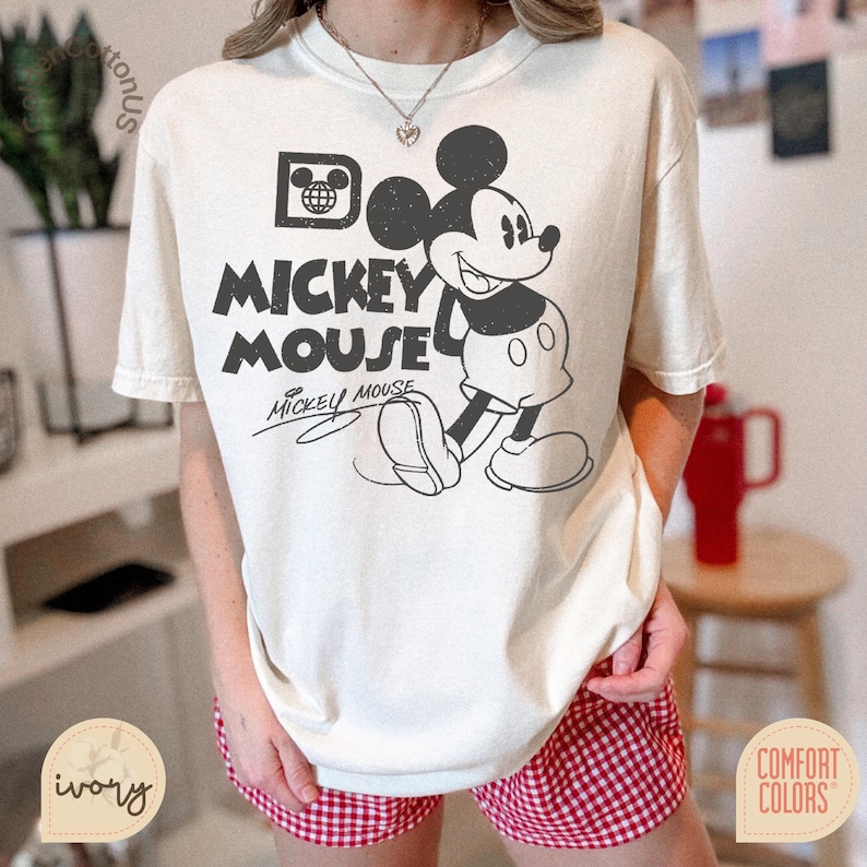 Puede incluir: Camiseta de color crema con un gran gr&aacute;fico en blanco y negro de Mickey Mouse. El dise&ntilde;o incluye las palabras "Mickey Mouse" en negrita, con la firma de Mickey debajo. La camiseta es de la marca Comfort Colors.