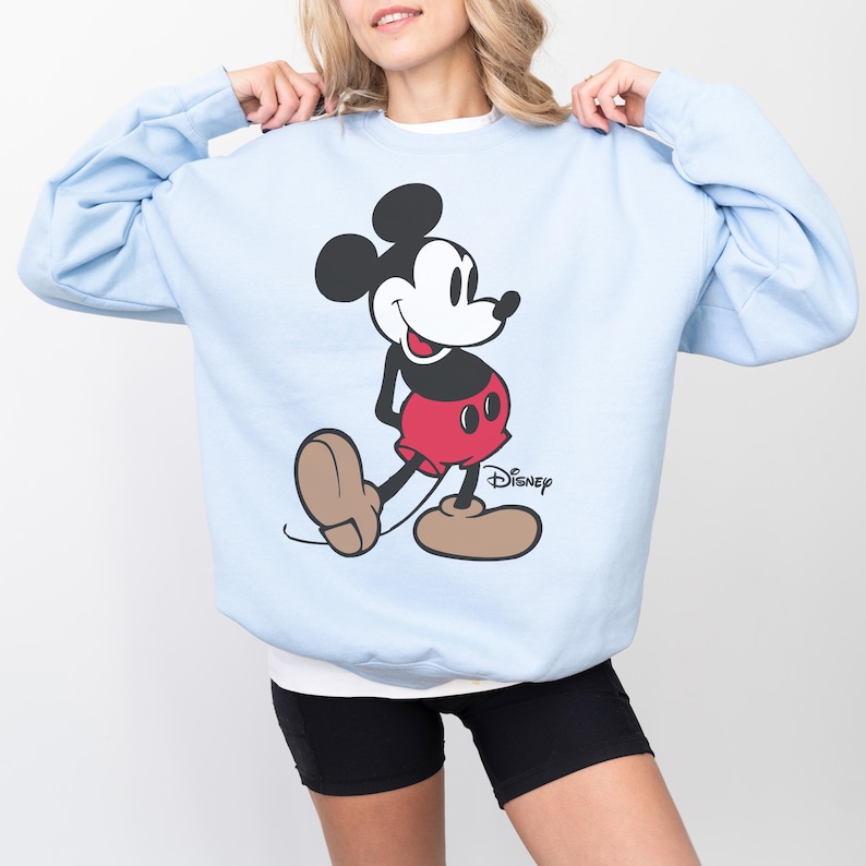 May include: Light blue sweatshirt featuring a large graphic of Mickey Mouse in his classic pose. The Disney character is depicted in black, red, and tan colors. The Disney logo is printed in black on the lower right side of the sweatshirt.