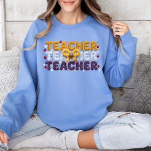 Puede incluir: Una sudadera de cuello redondo azul claro con un estampado gr&aacute;fico que repite la palabra TEACHER tres veces en diferentes patrones, incluyendo amarillo, papel rayado blanco y estampado de leopardo morado, con un lazo de l&aacute;piz amarillo y manzanas rojas.