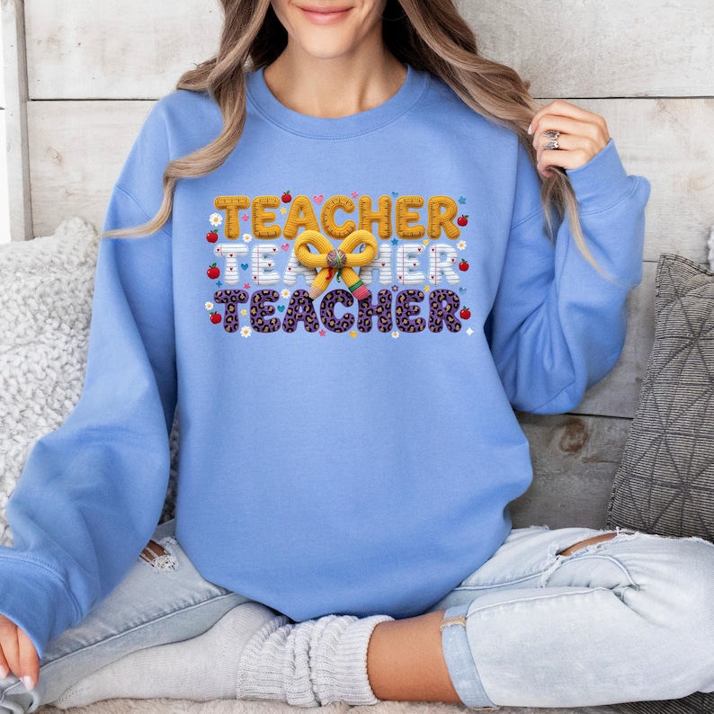 Puede incluir: Una sudadera de cuello redondo azul claro con un estampado gr&aacute;fico que repite la palabra TEACHER tres veces en diferentes patrones, incluyendo amarillo, papel rayado blanco y estampado de leopardo morado, con un lazo de l&aacute;piz amarillo y manzanas rojas.