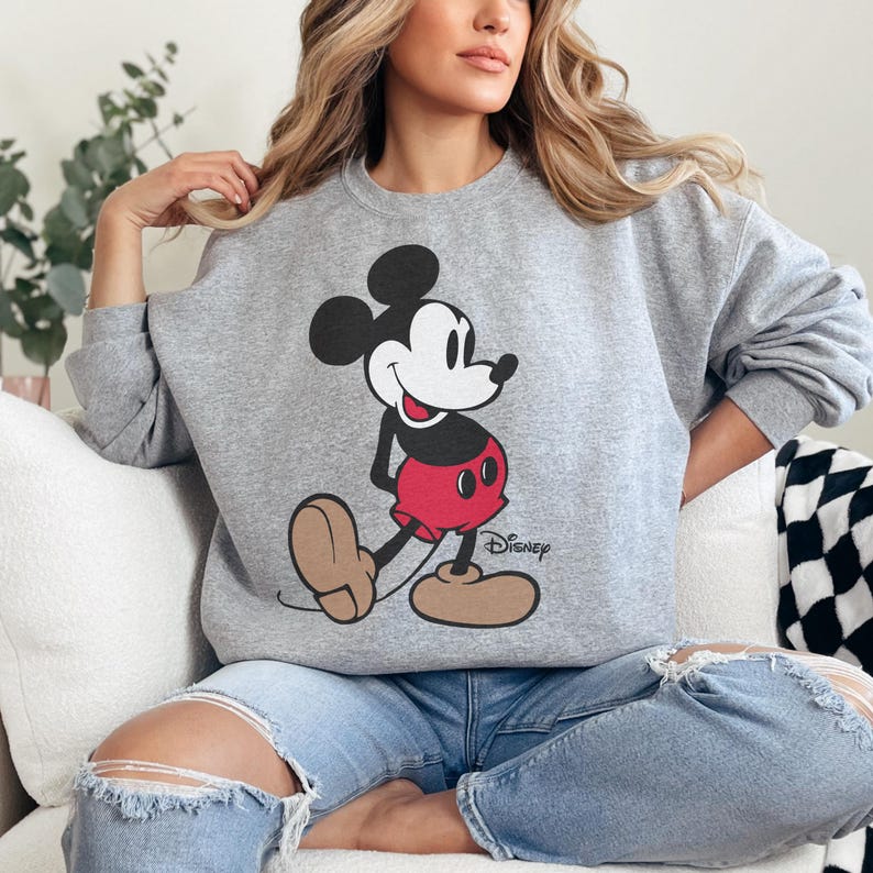 May include: A gray sweatshirt featuring a large graphic of Mickey Mouse in his classic pose. Mickey is depicted with a black head and ears, a white face, red shorts, and brown shoes. The Disney logo is printed below the character.