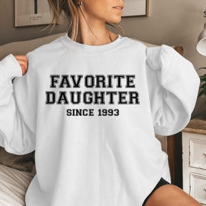 Peut inclure: Sweat-shirt blanc avec l'inscription "FAVORITE DAUGHTER SINCE 1993" en caract&egrave;res noirs gras. Le sweat-shirt est de style col rond, avec des manches longues. L'ensemble est d&eacute;contract&eacute; et confortable.
