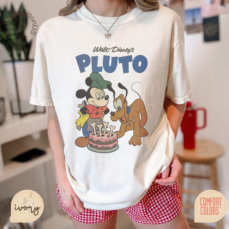 Puede incluir: Camiseta de color marfil con un gr&aacute;fico de Mickey Mouse y Pluto con un pastel. El texto "Walt Disney's PLUTO" est&aacute; impreso en azul sobre los personajes. La camiseta es de la marca Comfort Colors.