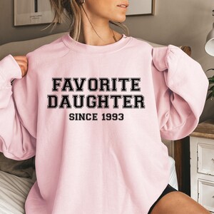 Peut inclure: Un sweat-shirt rose clair avec l'inscription "FAVORITE DAUGHTER SINCE 1993" en lettres noires. Le sweat-shirt &agrave; col rond semble &ecirc;tre fait d'une mati&egrave;re douce. L'image montre le sweat-shirt port&eacute;.