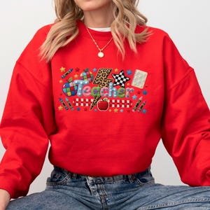 Puede incluir: Una sudadera roja de manga larga con un estampado gr&aacute;fico que muestra la palabra Teacher en letras coloridas. El dise&ntilde;o incluye un rayo con estampado de leopardo, una manzana roja, l&aacute;pices de colores, una estrella a cuadros y un cuaderno.