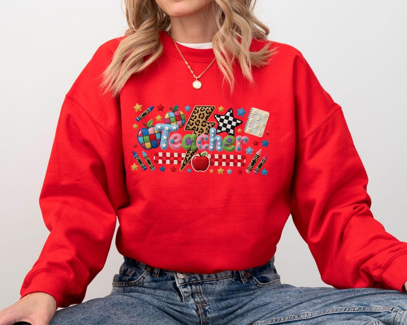 Puede incluir: Una sudadera roja de manga larga con un estampado gr&aacute;fico que muestra la palabra Teacher en letras coloridas. El dise&ntilde;o incluye un rayo con estampado de leopardo, una manzana roja, l&aacute;pices de colores, una estrella a cuadros y un cuaderno.
