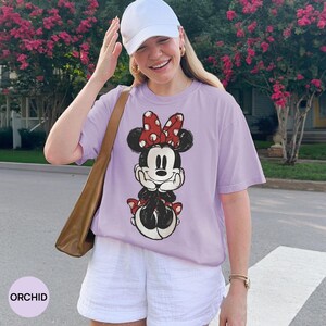 May include: Orchid-colored t-shirt featuring a Minnie Mouse graphic with a red polka dot bow. The shirt is paired with white shorts and a white baseball cap. A brown shoulder bag is also visible.