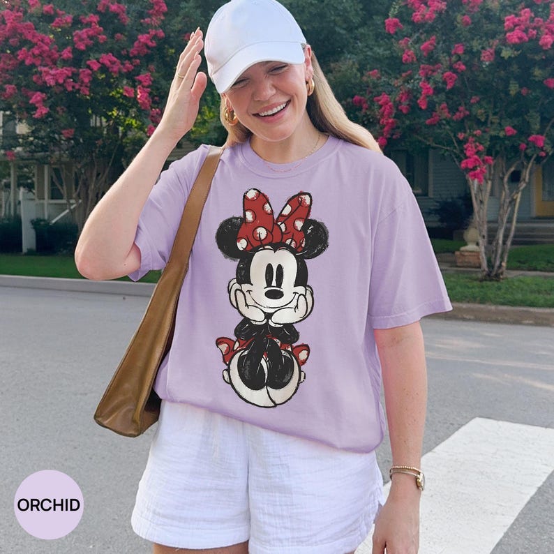 May include: Orchid-colored t-shirt featuring a Minnie Mouse graphic with a red polka dot bow. The shirt is paired with white shorts and a white baseball cap. A brown shoulder bag is also visible.