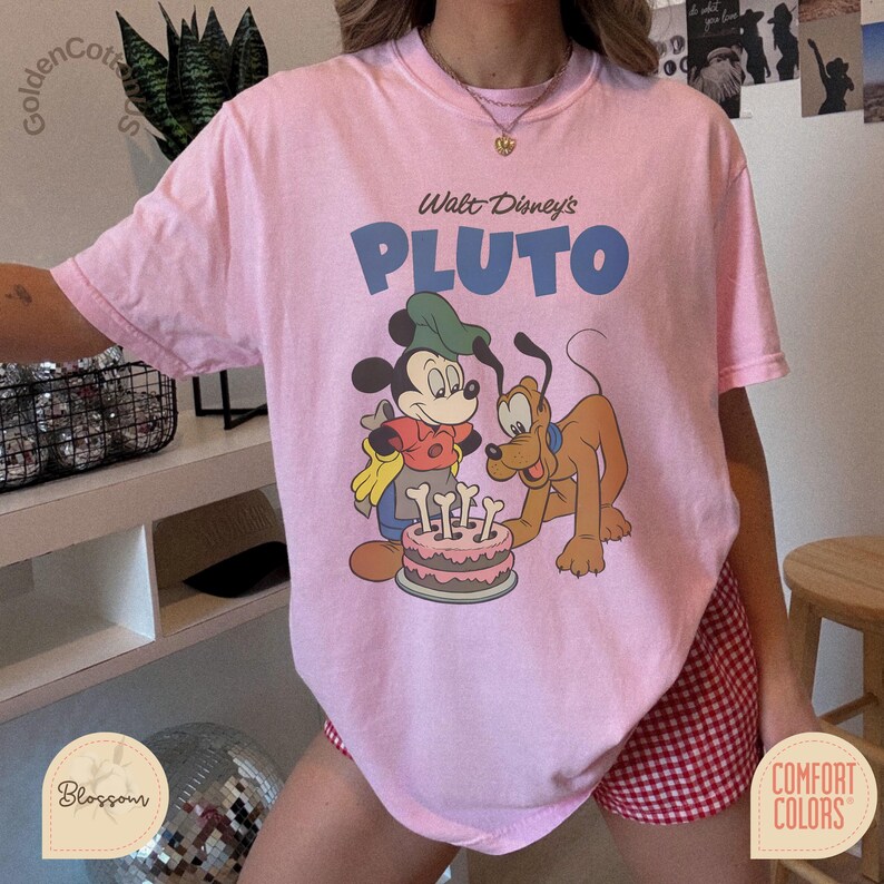 Puede incluir: Camiseta rosa claro con una ilustraci&oacute;n de Mickey Mouse y Pluto. El texto "Walt Disney's PLUTO" est&aacute; impreso en azul. La camiseta es de la marca Comfort Colors.