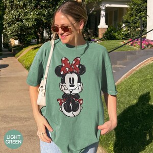 May include: Light green t-shirt featuring a Minnie Mouse graphic. The graphic shows Minnie Mouse in a seated pose, wearing a red bow with white polka dots. The shirt is a relaxed fit.