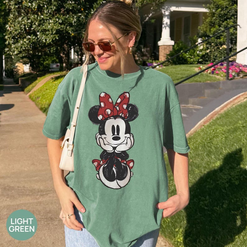 May include: Light green t-shirt featuring a Minnie Mouse graphic. The graphic shows Minnie Mouse in a seated pose, wearing a red bow with white polka dots. The shirt is a relaxed fit.