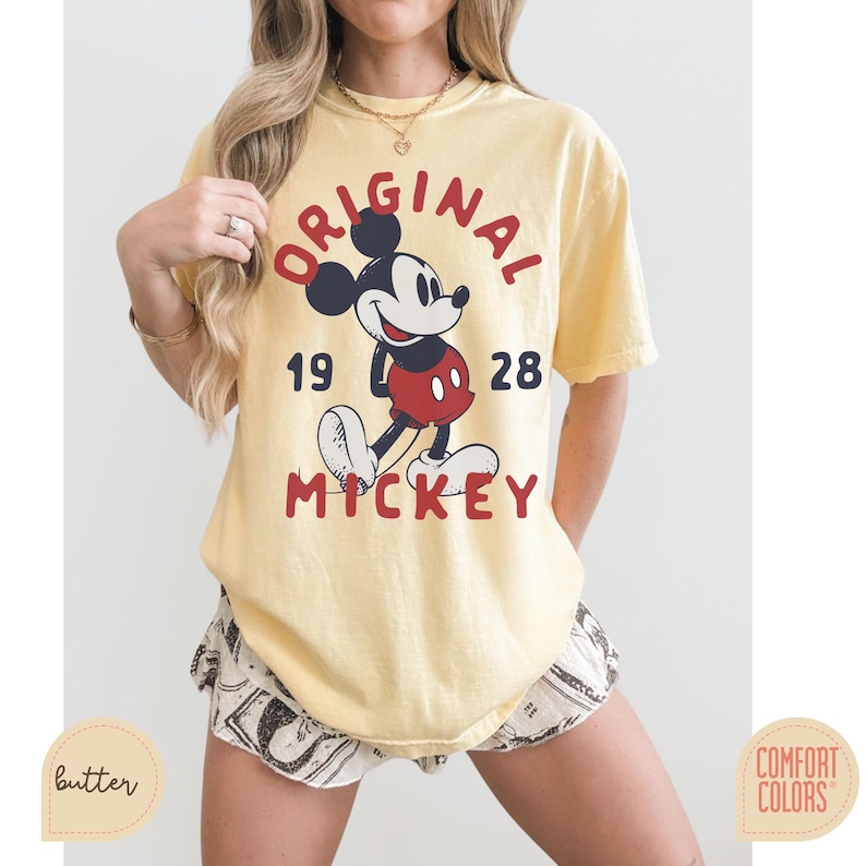 May include: Yellow t-shirt with a graphic of Mickey Mouse, the text "ORIGINAL MICKEY 1928" in red and black. The shirt is a Comfort Colors brand. The model is wearing patterned shorts.