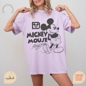 Puede incluir: Camiseta lila claro con un gr&aacute;fico en blanco y negro de Mickey Mouse. El dise&ntilde;o incluye las palabras "Mickey Mouse" y la firma de Mickey. La camiseta es de la marca Comfort Colors.