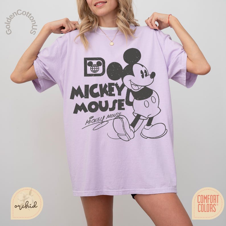Puede incluir: Camiseta lila claro con un gr&aacute;fico en blanco y negro de Mickey Mouse. El dise&ntilde;o incluye las palabras "Mickey Mouse" y la firma de Mickey. La camiseta es de la marca Comfort Colors.