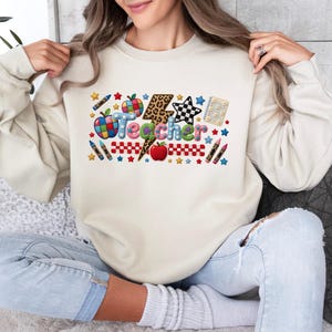 Puede incluir: Una sudadera de cuello redondo en color beige con un estampado gr&aacute;fico colorido que muestra la palabra Teacher en letras de burbuja. El dise&ntilde;o incluye un rayo con estampado de leopardo, manzanas rojas, crayones, una estrella a cuadros y estrellas.