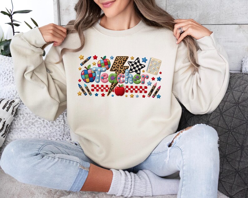 Puede incluir: Una sudadera de cuello redondo en color beige con un estampado gr&aacute;fico colorido que muestra la palabra Teacher en letras de burbuja. El dise&ntilde;o incluye un rayo con estampado de leopardo, manzanas rojas, crayones, una estrella a cuadros y estrellas.