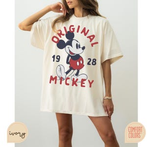 May include: Ivory-colored t-shirt featuring a graphic of Mickey Mouse with the text "ORIGINAL MICKEY 1928". The design includes red, navy, and black accents. The shirt is a Comfort Colors brand.