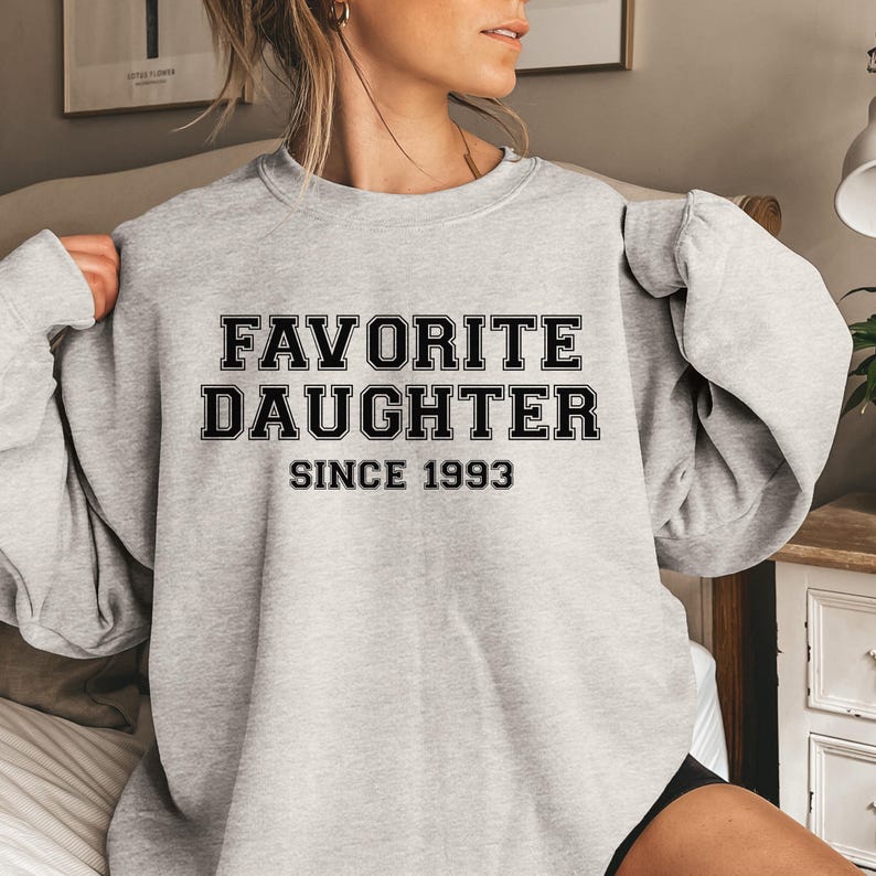 Peut inclure: Un sweat-shirt gris clair avec l'inscription "FAVORITE DAUGHTER SINCE 1993" en lettres noires. Le sweat-shirt est de style col rond et semble &ecirc;tre fait d'une mati&egrave;re douce.