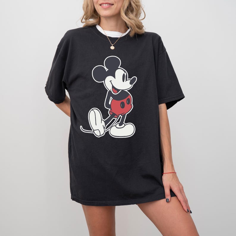 May include: Black t-shirt featuring a large graphic of Mickey Mouse in his classic pose. The cartoon character is depicted in black, red, and white. The shirt has a white collar and short sleeves.