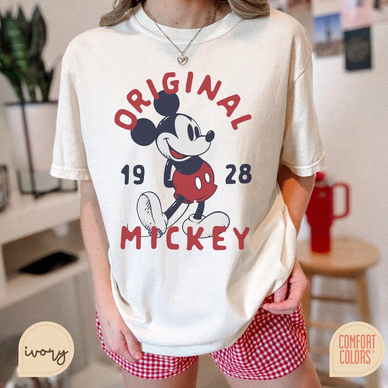May include: Ivory-colored t-shirt featuring a graphic of Mickey Mouse with the text "ORIGINAL MICKEY 1928" in red. The design includes Mickey in his classic red shorts and white gloves. The shirt is part of the Comfort Colors collection.
