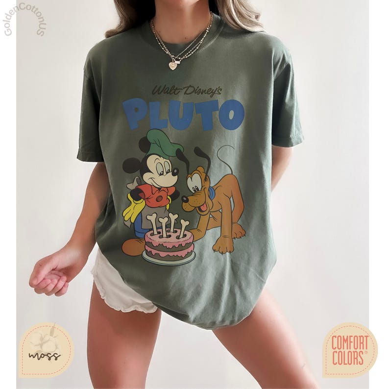 Puede incluir: Camiseta verde oliva con un gr&aacute;fico de Mickey Mouse y Pluto con una tarta de cumplea&ntilde;os. El texto "Walt Disney's PLUTO" es azul. La camiseta es de la marca Comfort Colors.