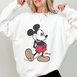 May include: White sweatshirt featuring a large graphic of Mickey Mouse. The cartoon character is depicted in his classic pose, wearing red shorts, black shoes, and black gloves. The Disney logo is printed on the bottom right.