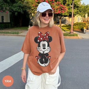 May include: Burnt orange t-shirt featuring a Minnie Mouse graphic. The cartoon character is depicted with a red and white polka dot bow, black ears, and a black and white outfit. The shirt is a relaxed fit.
