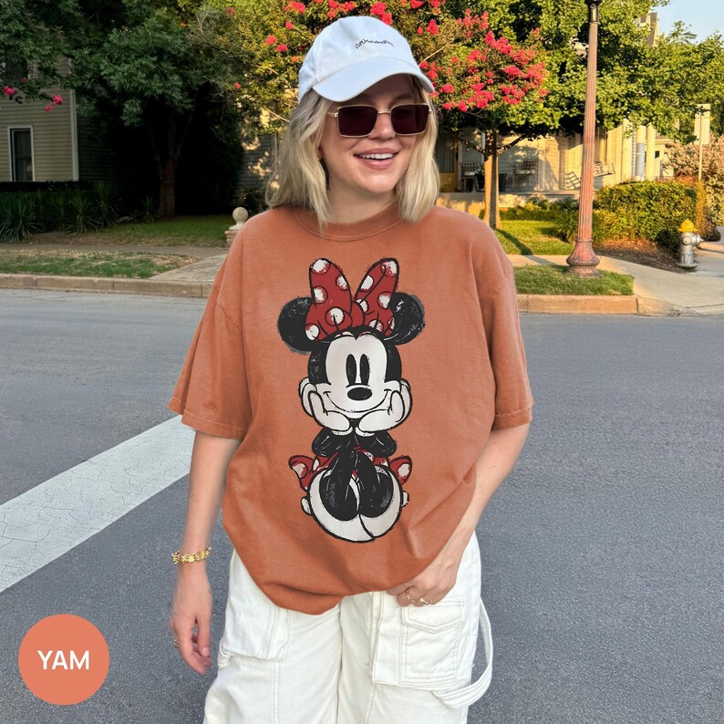 May include: Burnt orange t-shirt featuring a Minnie Mouse graphic. The cartoon character is depicted with a red and white polka dot bow, black ears, and a black and white outfit. The shirt is a relaxed fit.