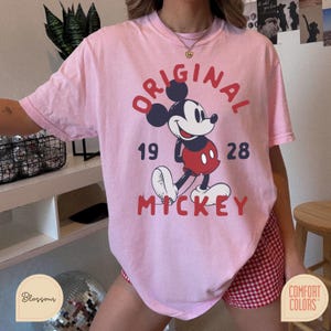 May include: A light pink t-shirt featuring a graphic of Mickey Mouse. The design includes the words "ORIGINAL MICKEY" in red, along with the numbers "19 28". The shirt is made of a soft material.