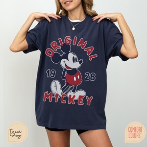 May include: Navy blue t-shirt featuring a classic Mickey Mouse graphic. The design includes the word "ORIGINAL" in red, the year "1928", and the name "MICKEY". The shirt is a Comfort Colors brand.