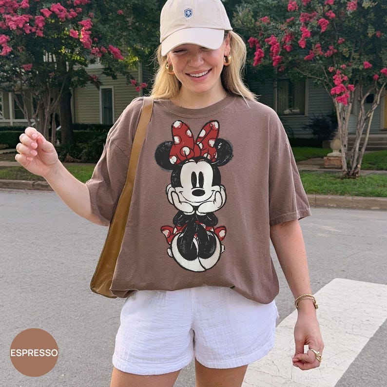 May include: Espresso-colored t-shirt featuring a Minnie Mouse graphic with a red bow and polka dots. The shirt is paired with white shorts and a beige baseball cap. A tan shoulder bag is also visible.