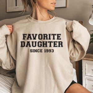 Peut inclure: Un sweat-shirt beige avec l'inscription "FAVORITE DAUGHTER SINCE 1993" en lettres noires. Le sweat-shirt a un col rond et des manches longues. L'image montre la partie sup&eacute;rieure du sweat-shirt.
