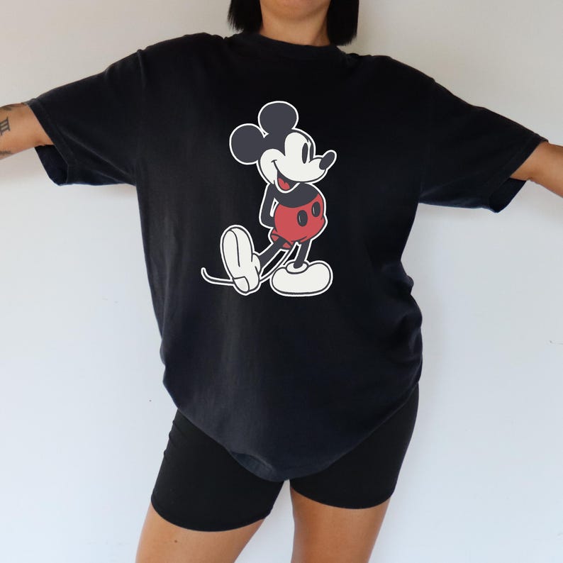 May include: Black t-shirt featuring a classic Mickey Mouse graphic. The cartoon character is depicted in his iconic pose, with a white face, black ears, red shorts, and white gloves and shoes. The shirt is a relaxed fit.