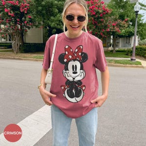 May include: Crimson-colored t-shirt featuring a Minnie Mouse graphic. The graphic shows Minnie Mouse in a classic pose, wearing a red bow with white polka dots. The shirt is paired with light blue jeans.