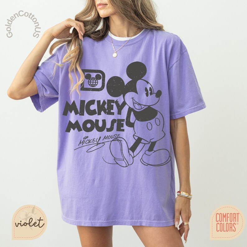 Puede incluir: Camiseta violeta Comfort Colors con un gran gr&aacute;fico negro de Mickey Mouse y las palabras "Mickey Mouse". La camiseta tiene mangas cortas y un corte relajado, perfecta para uso casual.