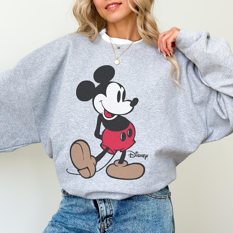 May include: A gray sweatshirt featuring a large graphic of Mickey Mouse in his classic pose. Mickey is depicted in black, red, and tan colors. The Disney logo is printed below the character.
