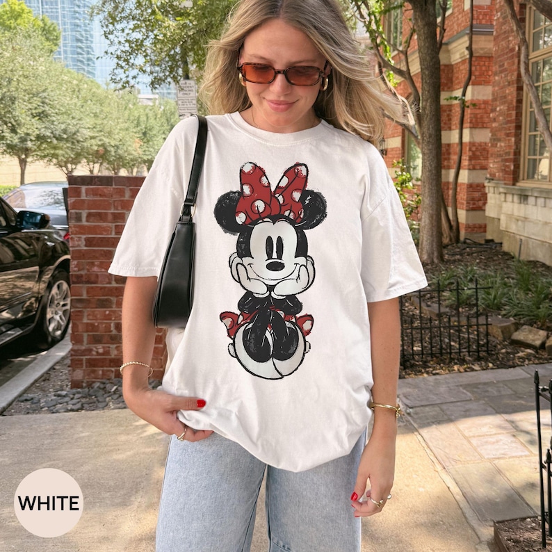 May include: White t-shirt featuring a Minnie Mouse graphic. The cartoon character is depicted in black, red, and white, with a large red polka-dot bow. The shirt is a casual style, suitable for everyday wear.