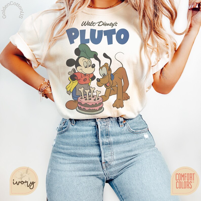 Puede incluir: Camiseta de color marfil con un dise&ntilde;o de Mickey Mouse y Pluto. El texto "Walt Disney's PLUTO" est&aacute; impreso en azul sobre los personajes. La camiseta es de la marca Comfort Colors.