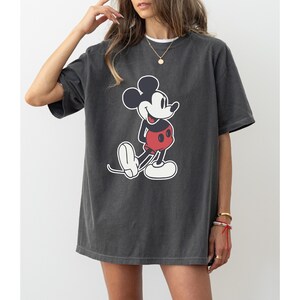 May include: Dark gray t-shirt featuring a classic Mickey Mouse graphic. The cartoon character is depicted in black, white, and red. The shirt has short sleeves and a relaxed fit, suitable for casual wear.